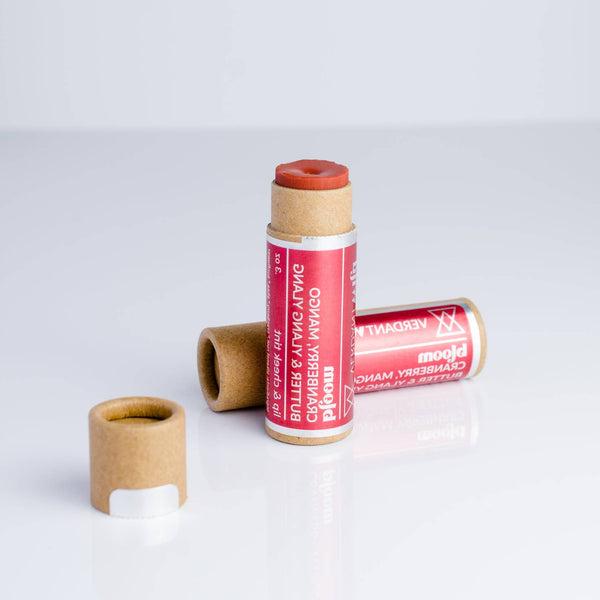 One cheek and lip balm standing with its cap off showing color of balm, and another one is placed on it side behind the standing one. Color of balm is a red orange color.